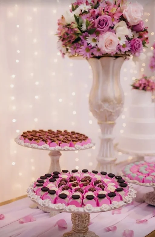 Mesa de doces de casamento montada pela Jana, com bombons em suportes, flores e bolo ao fundo