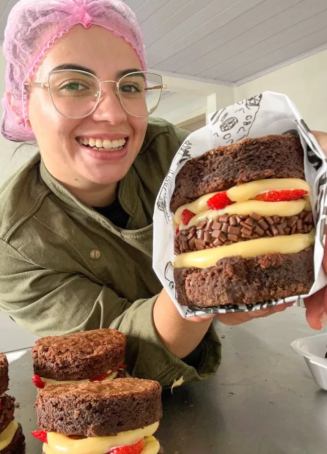 Jana sorrindo com um brownie recheado de morango nas mãos, na cozinha da doceria